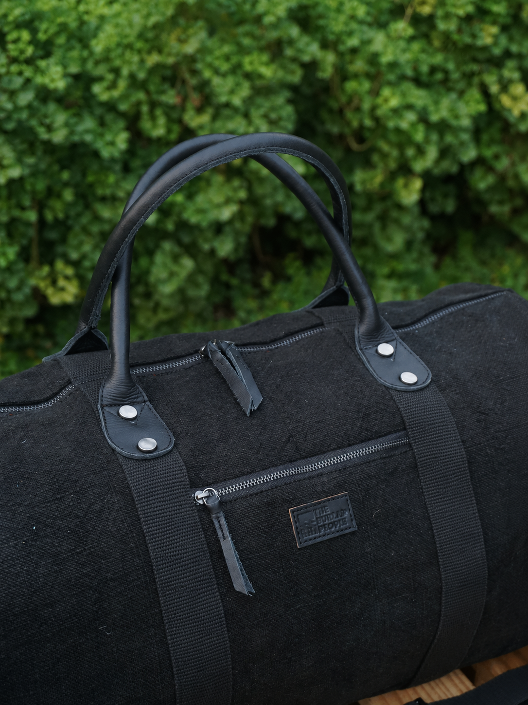 Black duffel bag with leather handles against a green bush background