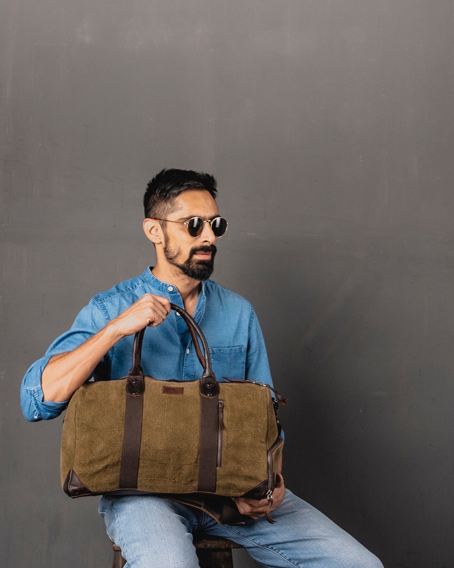 Brown Mini Duffel 2.0 in heavyweight burlap canvas held by top leather handles, worn by male model, showing front zip pocket and antique brass fittings.