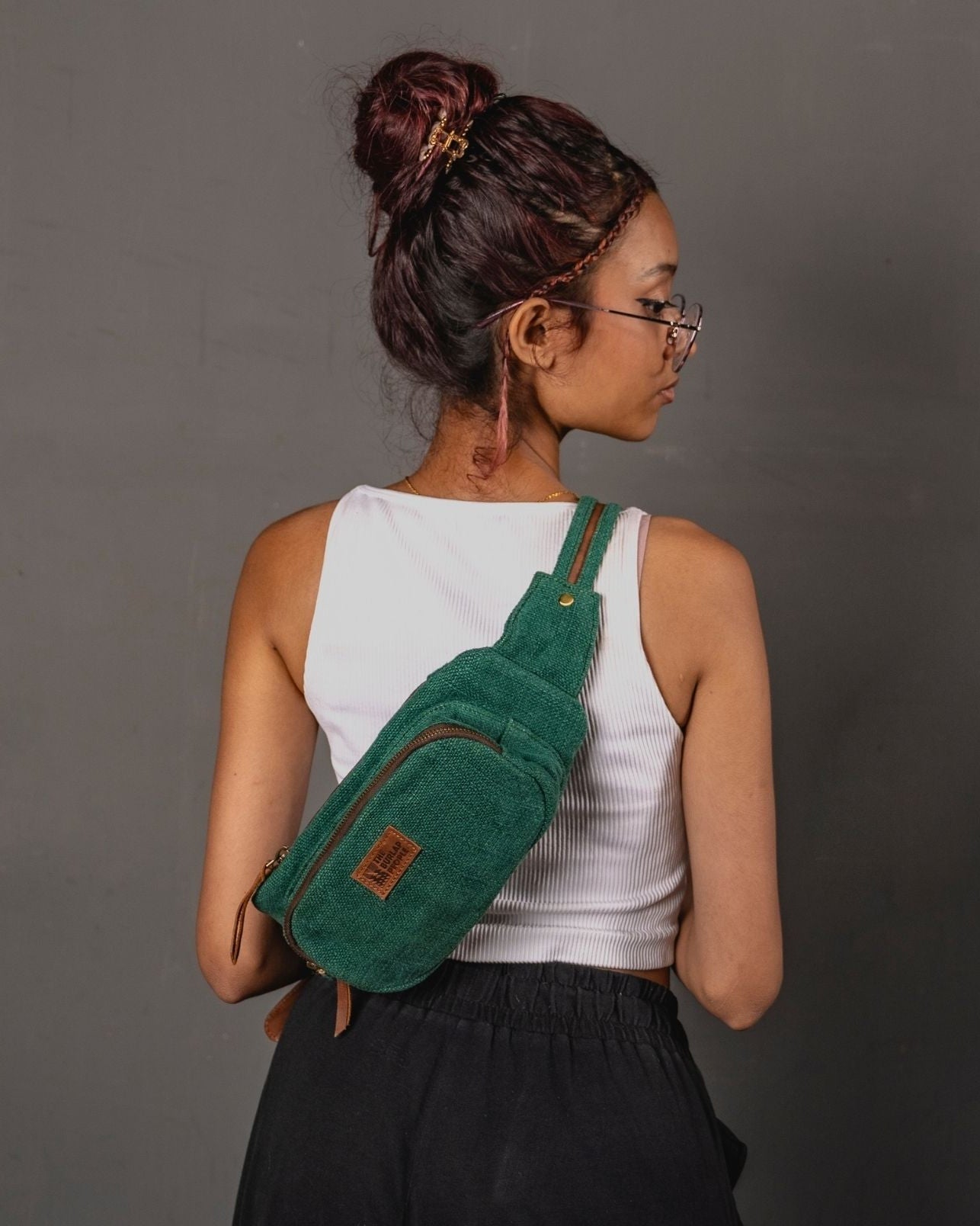 Woman wearing a bottle green burlap fanny pack on her back standing in front of a gray wall
